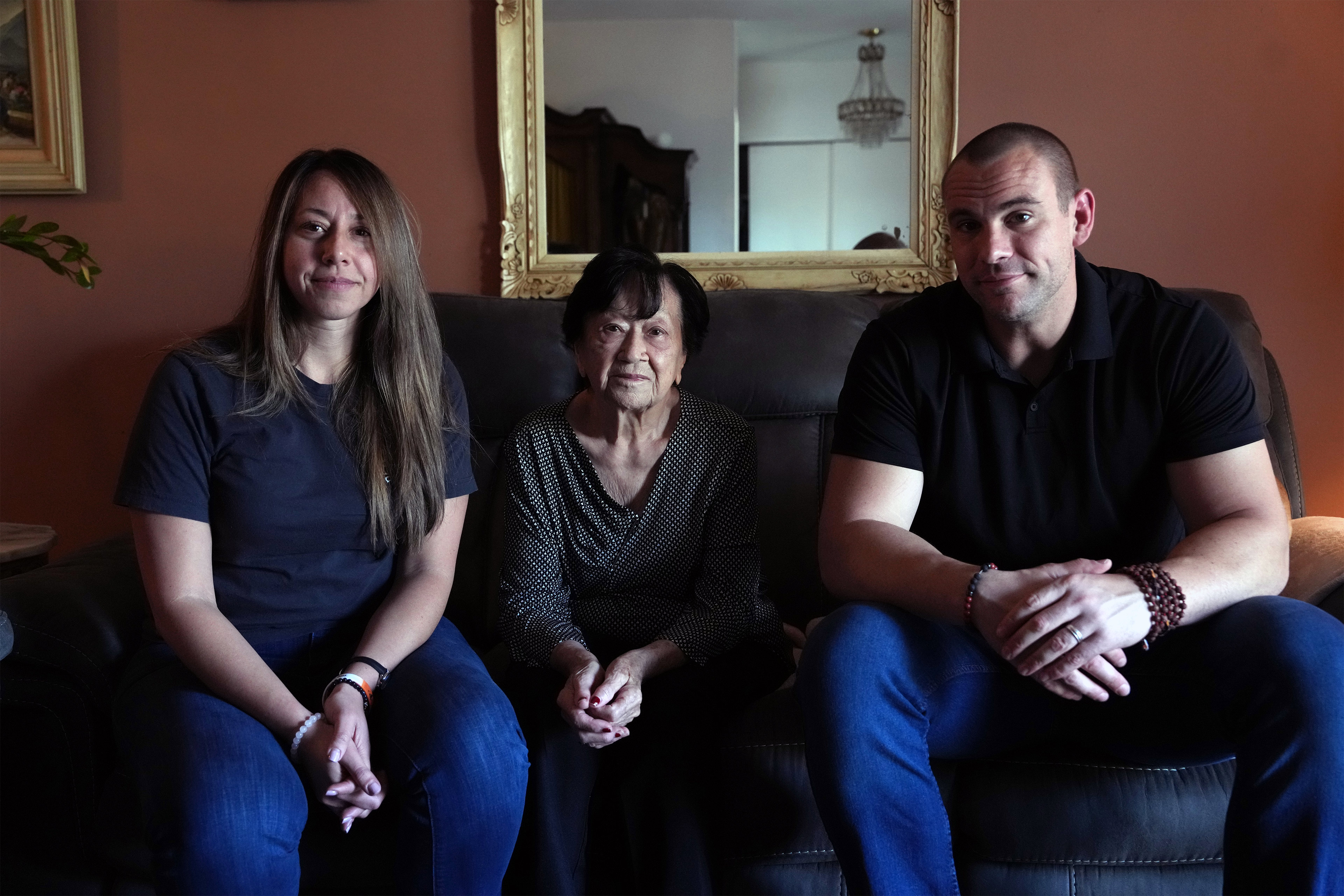 A photo of an older woman seated on a couch with her daughter and son-in-law sitting to her left and right.