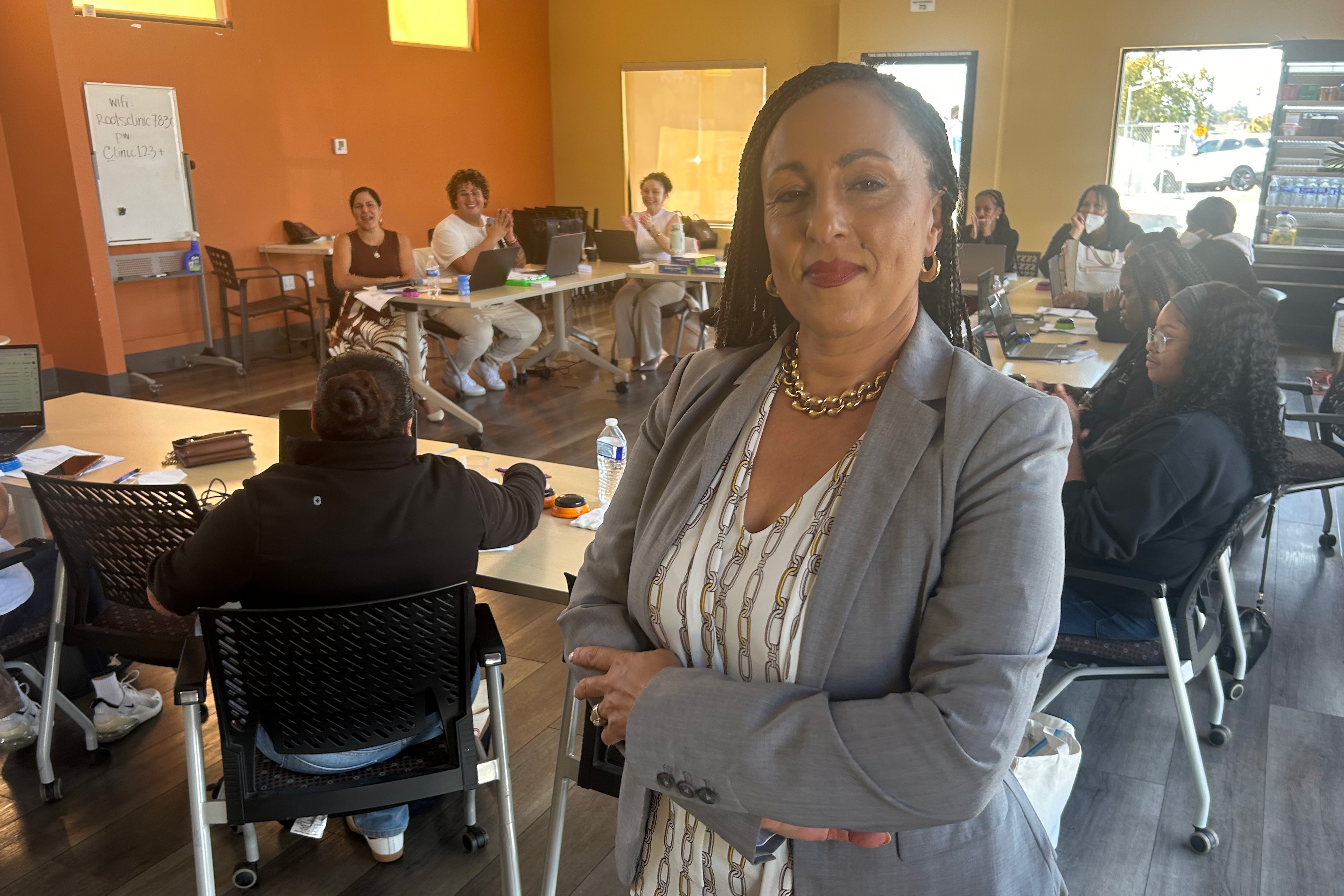 A woman in a suit stands in an office, several people seated at tables behind her