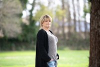 A woman with short blonde hair stands in a park outdoors. She is wearing a striped t-shirt, jeans, a black cardigan and a silver cross around her neck