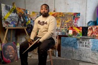 A photo shows Mykael Ash sitting in his studio, surrounded by his paintings behind him. He's holding a paintbrush and palette in his hands.