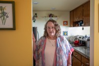 A woman in a pink tshirt and a paisley pink and blue cardigan stands in the doorway of a kitchen beside a yellow wall