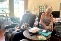 A woman wearing a sleeveless top, jeans, and glasses holds a drink and looks at her black-and-white dog, who sits in a recliner chair