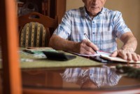 A senior man is sitting at a dining room table paying bills. He has a calculator in front of him.