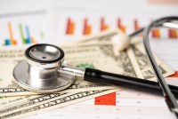 A stethoscope rests on top of a pile of U.S. dollar bills and finance report papers.