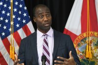 A man in a suit and tie speaks from behind a lectern. A U.S. flag and Florida state flag are behind him.