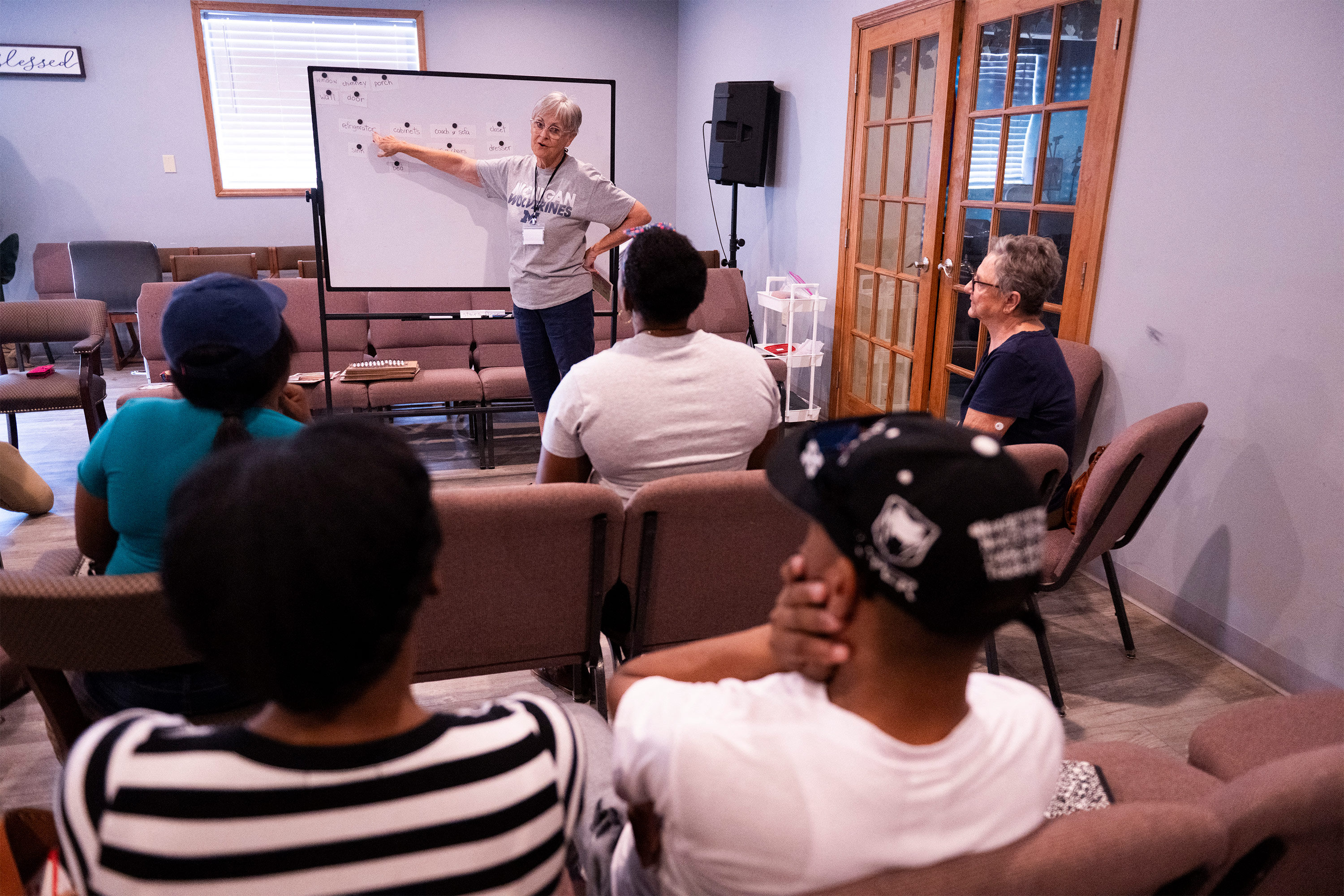 A photo of an older white woman teaching in front of a whiteboard to a class of adult Haitian students.