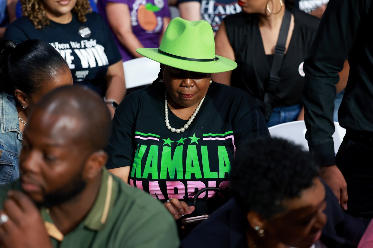 A photo of a Black women wearing a Kamala Harris shirt and a green hat that casts her eyes in shadow.