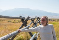 A man in a light colored long sleeve tshirt and glasses stands outside near a wooden fence