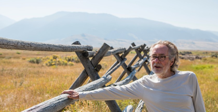 A man in a light colored long sleeve tshirt and glasses stands outside near a wooden fence