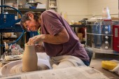 Denise Baker, a senior woman, works at a pottery wheel in a ceramics studio space.