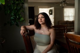 A woman with brown curly hair wearing a moss green top and gold necklace poses for a photo at a dining room table