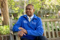 A photo of Lee Moultrie sitting on a bench outside in a blue pullover.