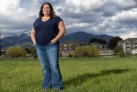 A woman in a dark blue t-shirt and jeans stands in a grassy field. There are buildings beyond the field and mountains farther in the distance.