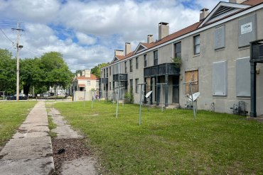 The back of a row of apartments where some of the windows are boarded up. A sidewalk runs parallel to the building.